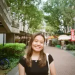 Smiling young woman standing outdoors in a city street with trees and shops.