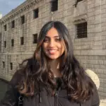 Smiling young woman with long dark hair in front of historic stone building.