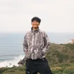 Young man smiling outdoors near the coast with hills in the background.