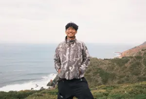 Young man smiling outdoors near the coast with hills in the background.