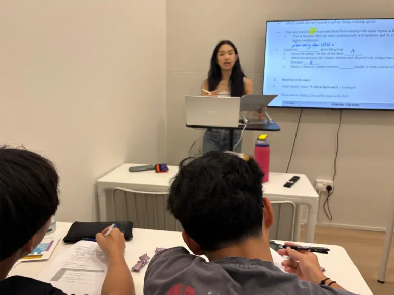 Young woman teaching chemistry in a classroom with students taking notes.