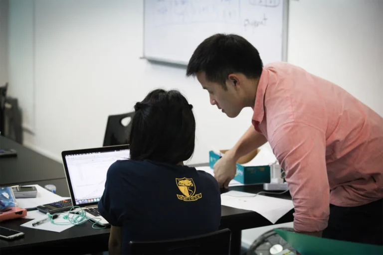 Teacher assisting student with computer work in classroom setting.
