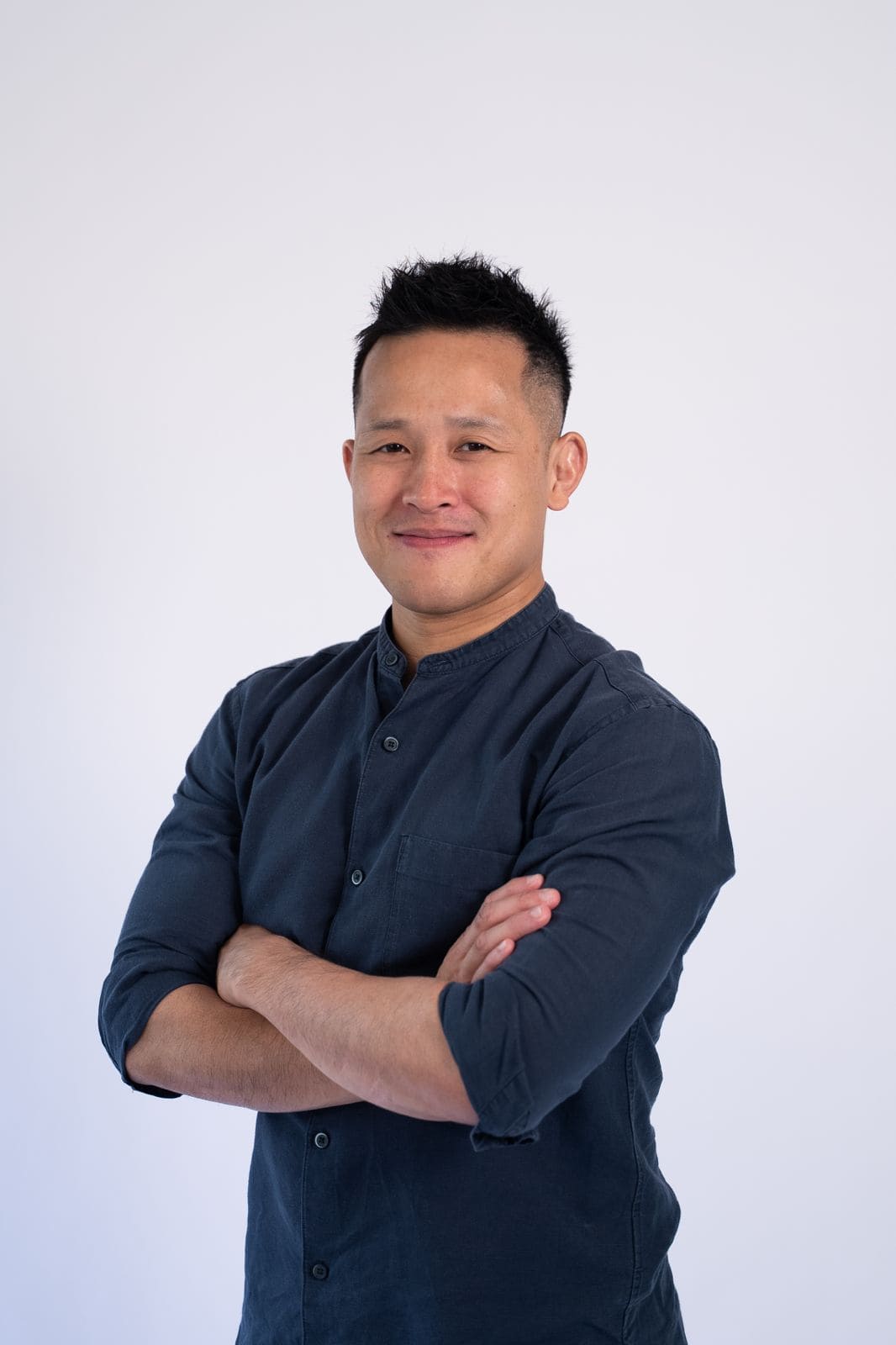 Confident Asian man in navy shirt with arms crossed against white background.
