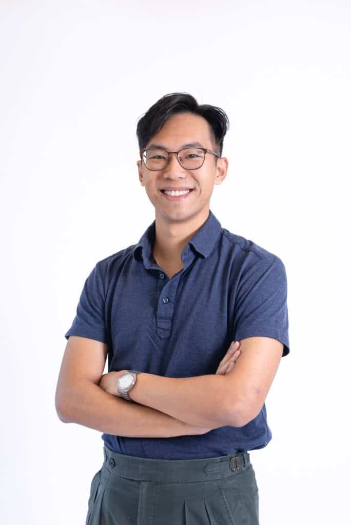 Young man with glasses smiling, arms crossed, in a blue shirt and grey trousers.