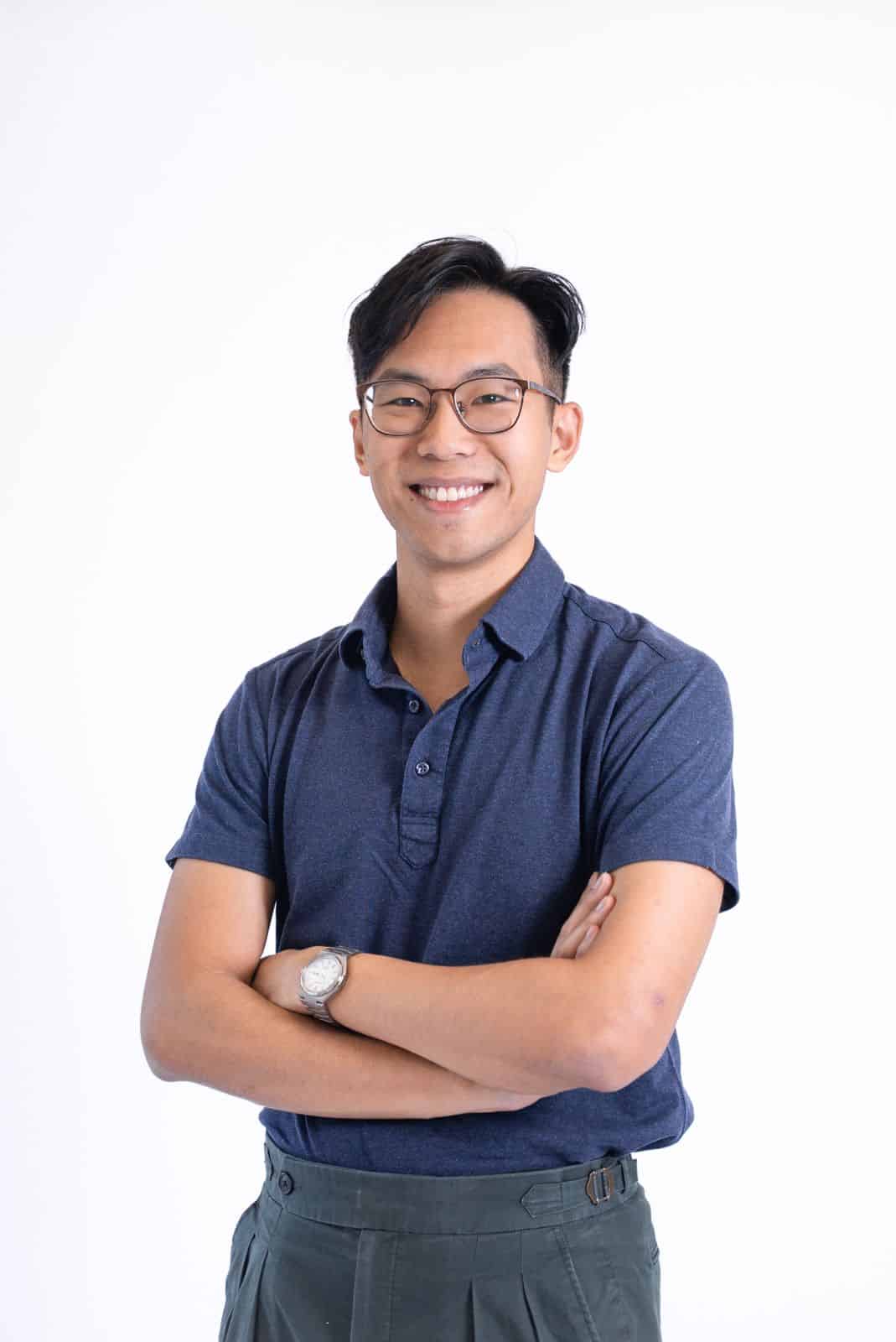 Young man with glasses smiling, arms crossed, in a blue shirt and grey trousers.