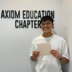 Young man smiling at Axiom Education Chapter sign holding a paper.