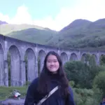 Young woman smiling outdoors near a historic stone viaduct in a lush green valley.