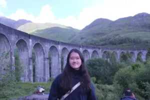 Young woman smiling outdoors near a historic stone viaduct in a lush green valley.
