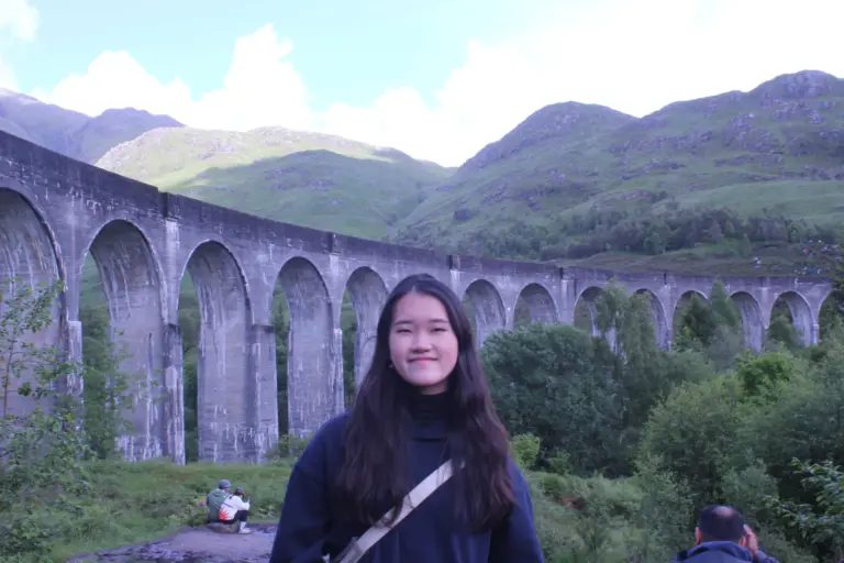 Young woman smiling outdoors near a historic stone viaduct in a lush green valley.