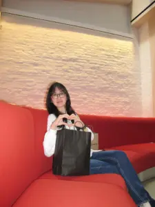 Young woman with glasses holding a black shopping bag on a red sofa.