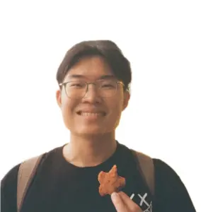 Young man with glasses smiling, holding a small snack, wearing a black T-shirt and backpack.
